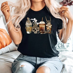 K&ouml;nnte beinhalten: Schwarzes T-Shirt mit einem Motiv von drei Kaffeetassen, die von Skeletten gehalten werden. Die Tassen sind mit Kaffee und Schlagsahne gef&uuml;llt.