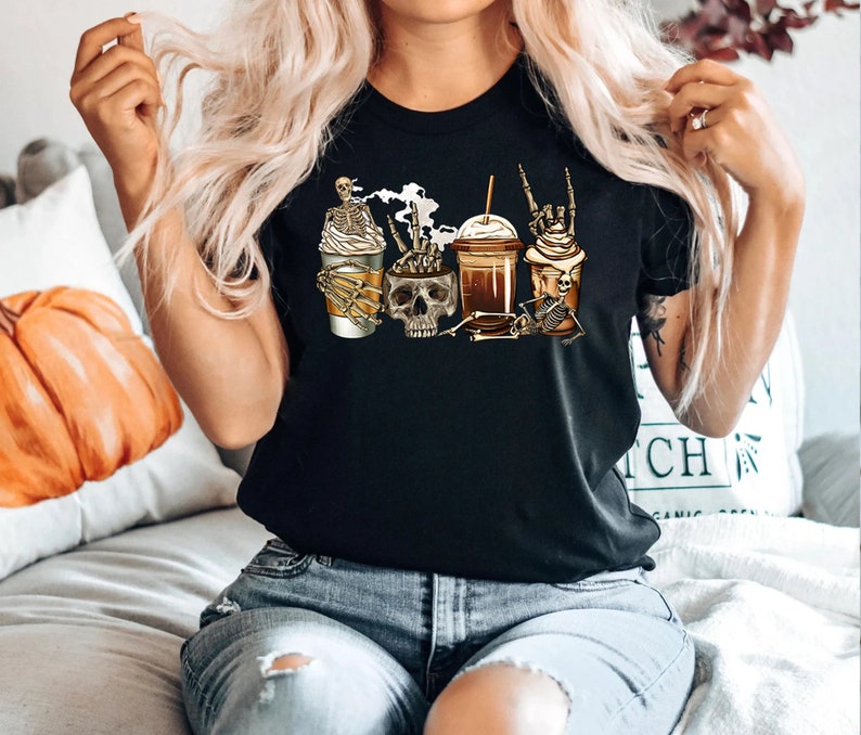 K&ouml;nnte beinhalten: Schwarzes T-Shirt mit einem Motiv von drei Kaffeetassen, die von Skeletten gehalten werden. Die Tassen sind mit Kaffee und Schlagsahne gef&uuml;llt.