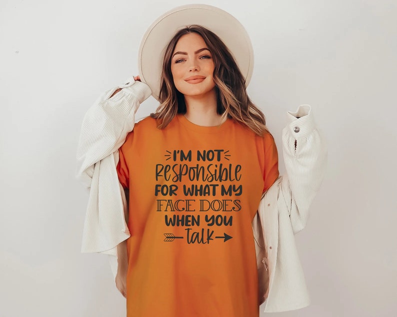 K&ouml;nnte beinhalten: Orangefarbenes T-Shirt mit schwarzem Text, der "Ich bin nicht verantwortlich f&uuml;r das, was mein Gesicht tut, wenn du sprichst" lautet, mit Pfeilen auf beiden Seiten des Textes.