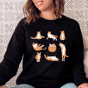 K&ouml;nnte beinhalten: Schwarzes Sweatshirt mit einem Aufdruck von orangefarbenen und wei&szlig;en Katzen in verschiedenen Yoga-Posen. Die Katzen machen Posen wie Downward Dog, Cat Cow und Child's Pose.