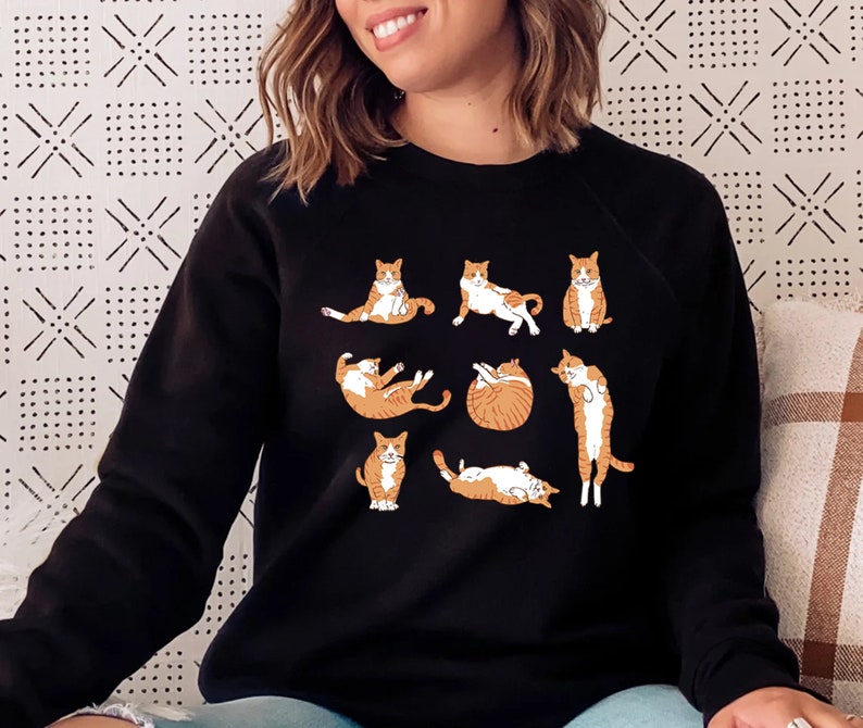 K&ouml;nnte beinhalten: Schwarzes Sweatshirt mit einem Aufdruck von orangefarbenen und wei&szlig;en Katzen in verschiedenen Yoga-Posen. Die Katzen machen Posen wie Downward Dog, Cat Cow und Child's Pose.