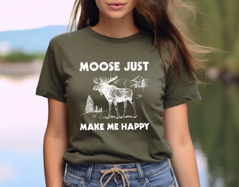 May include: Olive green t-shirt with a white graphic of a moose, trees, and mountains. The text "MOOSE JUST MAKE ME HAPPY" is printed above and below the illustration. The shirt is made of a soft material.