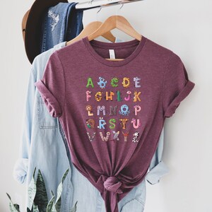 May include: A maroon t-shirt with a colorful alphabet printed on the front. Each letter is decorated with a different animal illustration.