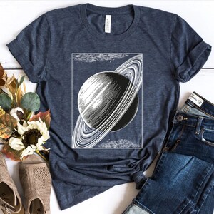 May include: A navy blue t-shirt with a vintage illustration of Saturn with its rings. The shirt is laid flat on a white wooden surface with a pair of brown sandals and a pair of blue jeans.
