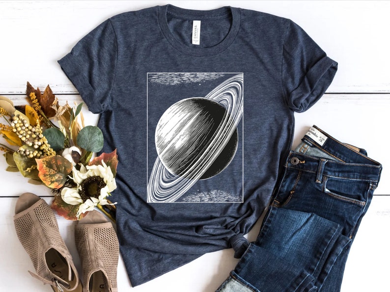 May include: A navy blue t-shirt with a vintage illustration of Saturn with its rings. The shirt is laid flat on a white wooden surface with a pair of brown sandals and a pair of blue jeans.