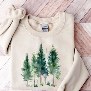 Puede incluir: Una sudadera beige con un estampado de acuarela de una escena de bosque. Los &aacute;rboles son verdes y el fondo es un lavado ligero de verde y azul. La sudadera tiene cuello redondo y mangas largas.