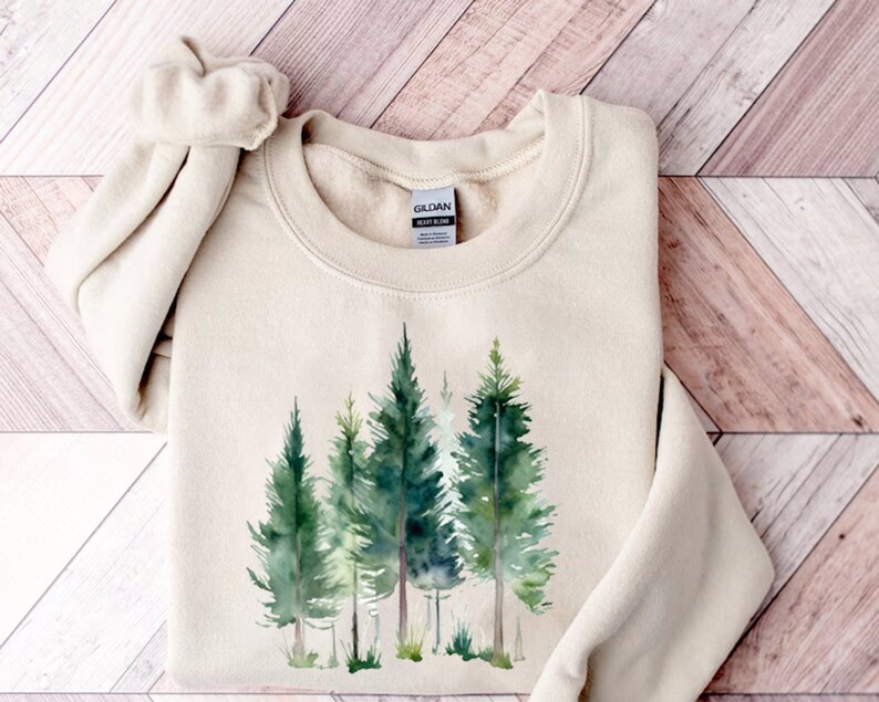 Puede incluir: Una sudadera beige con un estampado de acuarela de una escena de bosque. Los &aacute;rboles son verdes y el fondo es un lavado ligero de verde y azul. La sudadera tiene cuello redondo y mangas largas.