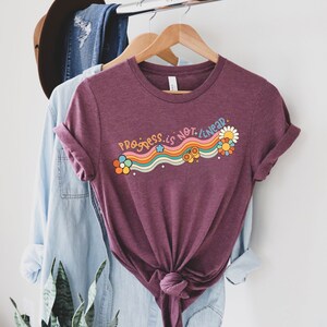 K&ouml;nnte beinhalten: Ein maroonfarbenes T-Shirt mit dem Text "Progress is not linear" in Regenbogen-Schrift mit Blumen und Sternen.