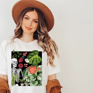 May include: White t-shirt with a black square graphic featuring a skeleton holding a watering can with the text "You make me feel alive". The graphic also includes green and red flowers, butterflies, and various plants.