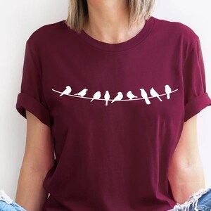 May include: A maroon t-shirt with a white graphic of a line of birds perched on a wire.