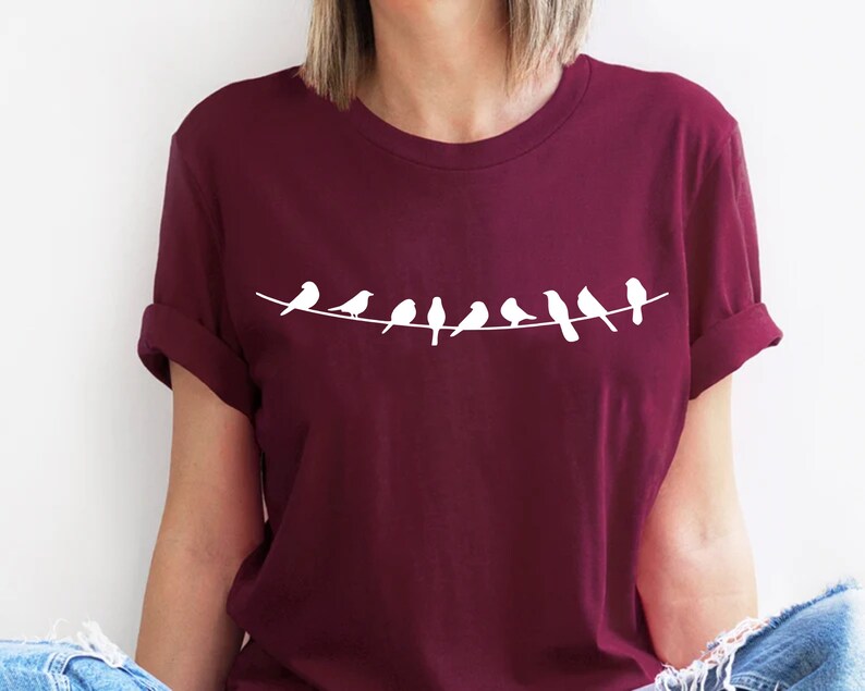 May include: A maroon t-shirt with a white graphic of a line of birds perched on a wire.
