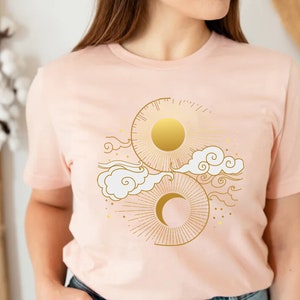 May include: A pink t-shirt with a gold sun and moon design. The design features a sun with rays, a crescent moon, and stylized clouds.