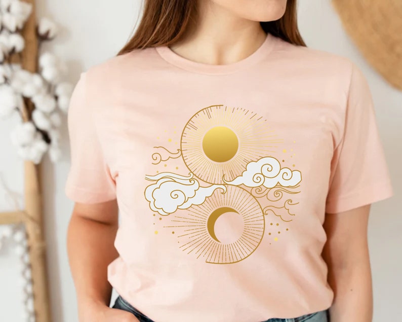May include: A pink t-shirt with a gold sun and moon design. The design features a sun with rays, a crescent moon, and stylized clouds.