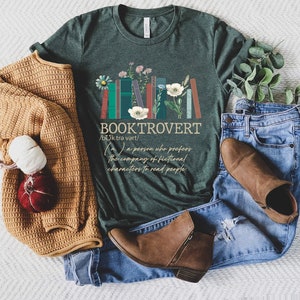 Puede incluir: Una camiseta verde con un gr&aacute;fico de libros apilados con flores y el texto "BOOKTROVERT /bok trə vart/ (m.) a person who prefers the company of fictional characters to read people."