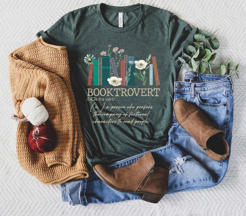 Puede incluir: Una camiseta verde con un gr&aacute;fico de libros apilados con flores y el texto "BOOKTROVERT /bok trə vart/ (m.) a person who prefers the company of fictional characters to read people."