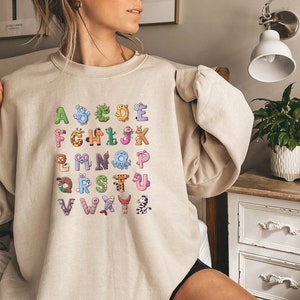 May include: A beige sweatshirt with a graphic print of the alphabet. Each letter is decorated with a different animal or creature, such as a giraffe, an octopus, and a zebra.