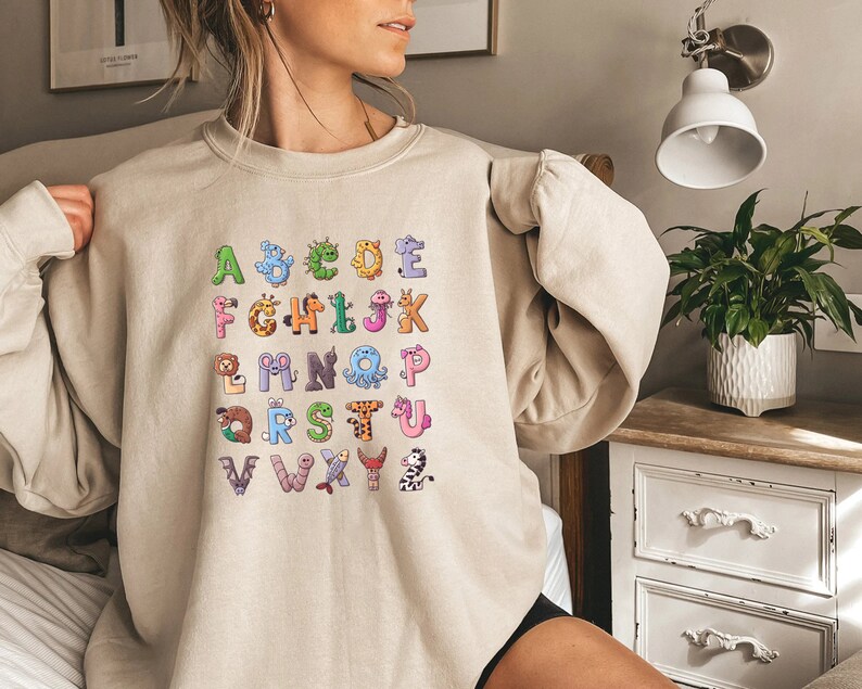 May include: A beige sweatshirt with a graphic print of the alphabet. Each letter is decorated with a different animal or creature, such as a giraffe, an octopus, and a zebra.