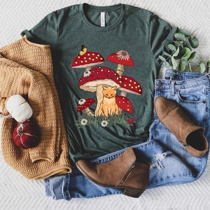 May include: A green t-shirt with a graphic of a cat sitting under a cluster of red and white mushrooms. The mushrooms have white spots and a butterfly is perched on one of them.