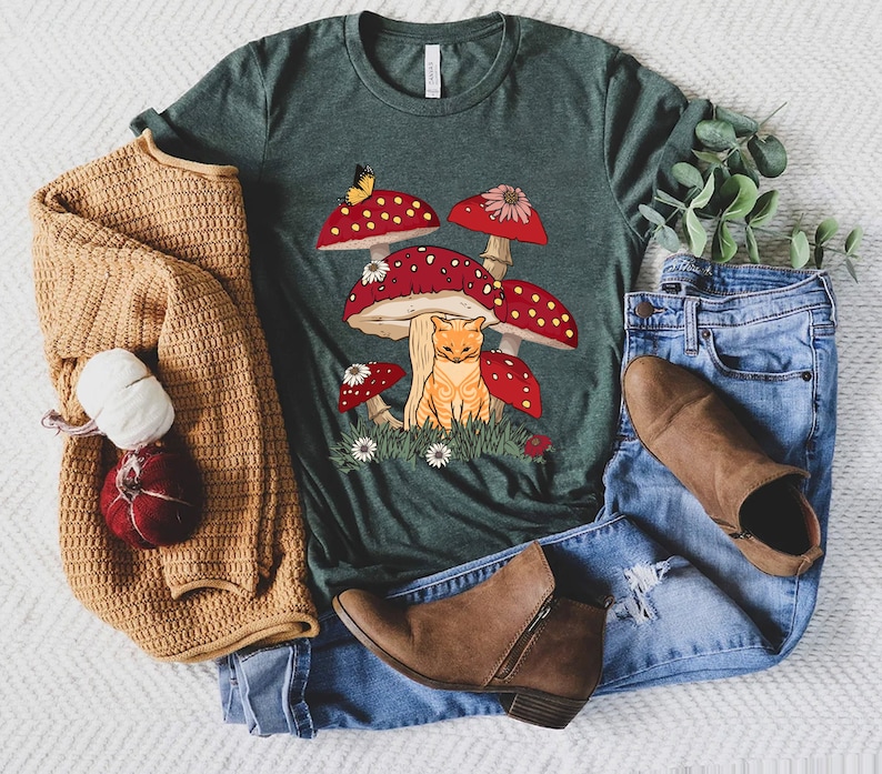 May include: A green t-shirt with a graphic of a cat sitting under a cluster of red and white mushrooms. The mushrooms have white spots and a butterfly is perched on one of them.