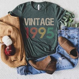 May include: A dark green t-shirt with the text "Vintage 1995" in rainbow colors. The shirt is laid out on a white surface with a brown sweater, blue jeans, and brown boots.