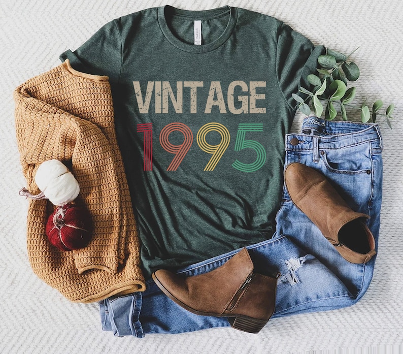May include: A dark green t-shirt with the text "Vintage 1995" in rainbow colors. The shirt is laid out on a white surface with a brown sweater, blue jeans, and brown boots.