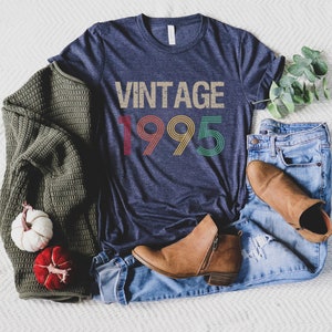 May include: A navy blue t-shirt with the text "Vintage 1995" in rainbow colors. The shirt is laid out on a white surface with a pair of brown ankle boots, blue jeans, and a green sweater.