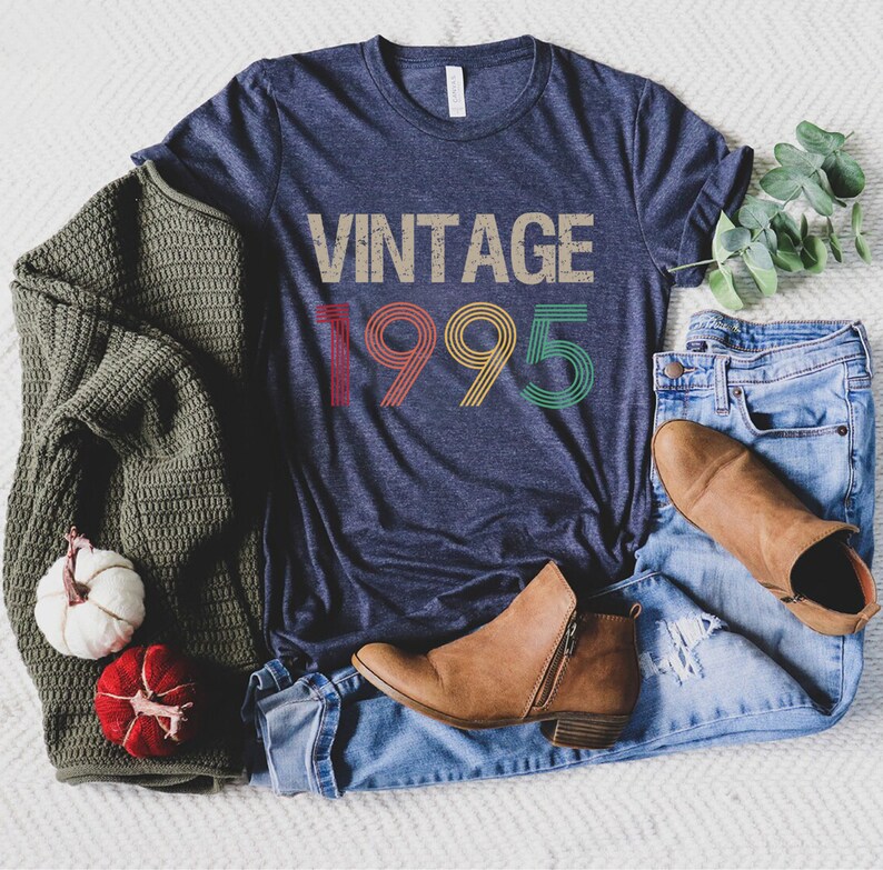 May include: A navy blue t-shirt with the text "Vintage 1995" in rainbow colors. The shirt is laid out on a white surface with a pair of brown ankle boots, blue jeans, and a green sweater.