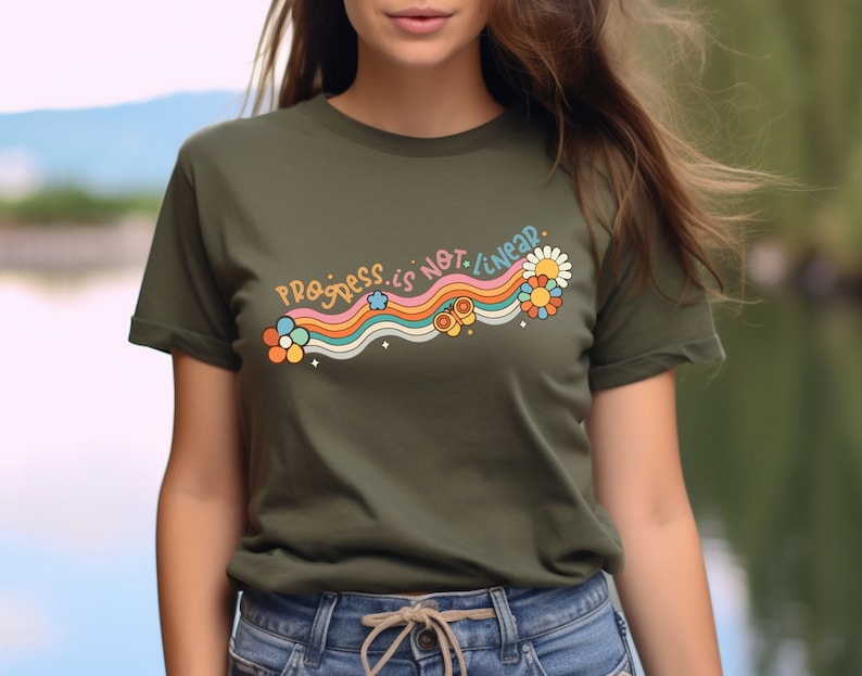 K&ouml;nnte beinhalten: Ein gr&uuml;nes T-Shirt mit einem Regenbogen- und Blumendesign. Der Text auf dem Shirt lautet "Progress is not linear."
