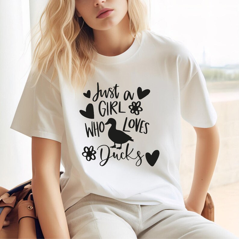 May include: White t-shirt with the text "Just a GIRL WHO LOVES Ducks" in black. The design includes hearts, flowers, and a silhouette of a duck. The shirt is a casual, short-sleeved style.