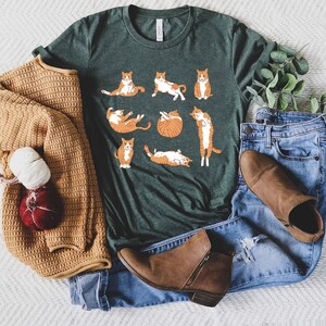 May include: A dark green t-shirt featuring a playful design of orange and white cats in various poses. The shirt is paired with blue jeans, brown ankle boots, and a tan sweater. The overall aesthetic is casual and comfortable.