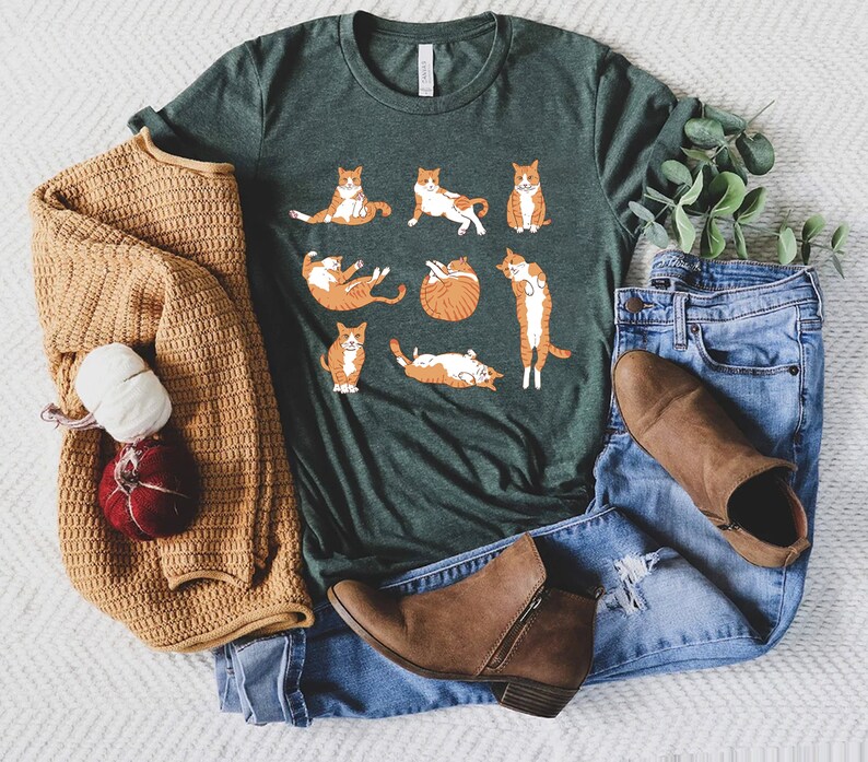 May include: A dark green t-shirt featuring a playful design of orange and white cats in various poses. The shirt is paired with blue jeans, brown ankle boots, and a tan sweater. The overall aesthetic is casual and comfortable.