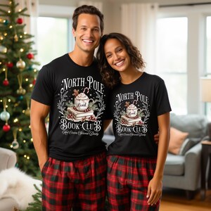 K&ouml;nnte beinhalten: Zwei Personen tragen schwarze T-Shirts mit einem weihnachtlichen Design, auf dem "North Pole Book Club" und "Hot Cocoa Served Daily" steht.