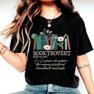 Puede incluir: Camiseta negra con un gr&aacute;fico de una pila de libros con flores y el texto "BOOKTROVERT /bUk trə vərt/ (n.) a person who prefers the company of fictional characters to read people."