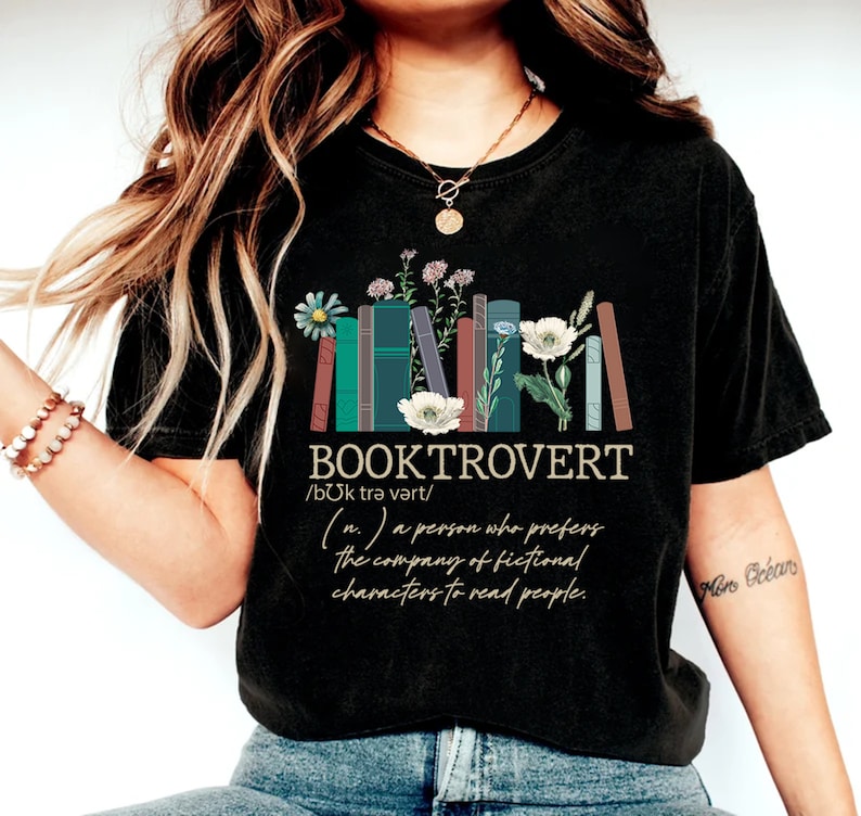 Puede incluir: Camiseta negra con un gr&aacute;fico de una pila de libros con flores y el texto "BOOKTROVERT /bUk trə vərt/ (n.) a person who prefers the company of fictional characters to read people."