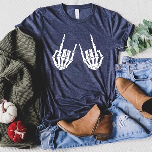 May include: A navy blue t-shirt with a white graphic of two skeleton hands giving the middle finger. The shirt is laying on a white surface with a pair of jeans and brown boots.