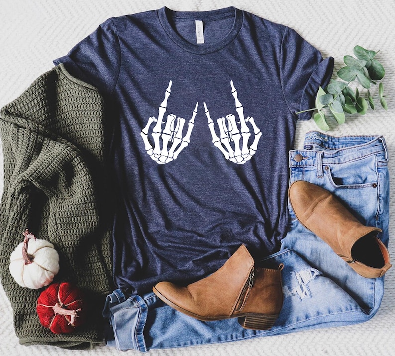 May include: A navy blue t-shirt with a white graphic of two skeleton hands giving the middle finger. The shirt is laying on a white surface with a pair of jeans and brown boots.