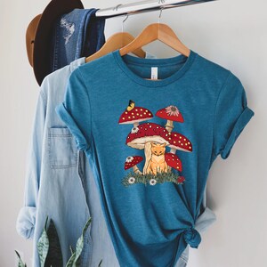 May include: A teal blue t-shirt with a graphic of a cat sitting under a cluster of red and white mushrooms. A yellow butterfly is flying above the mushrooms.