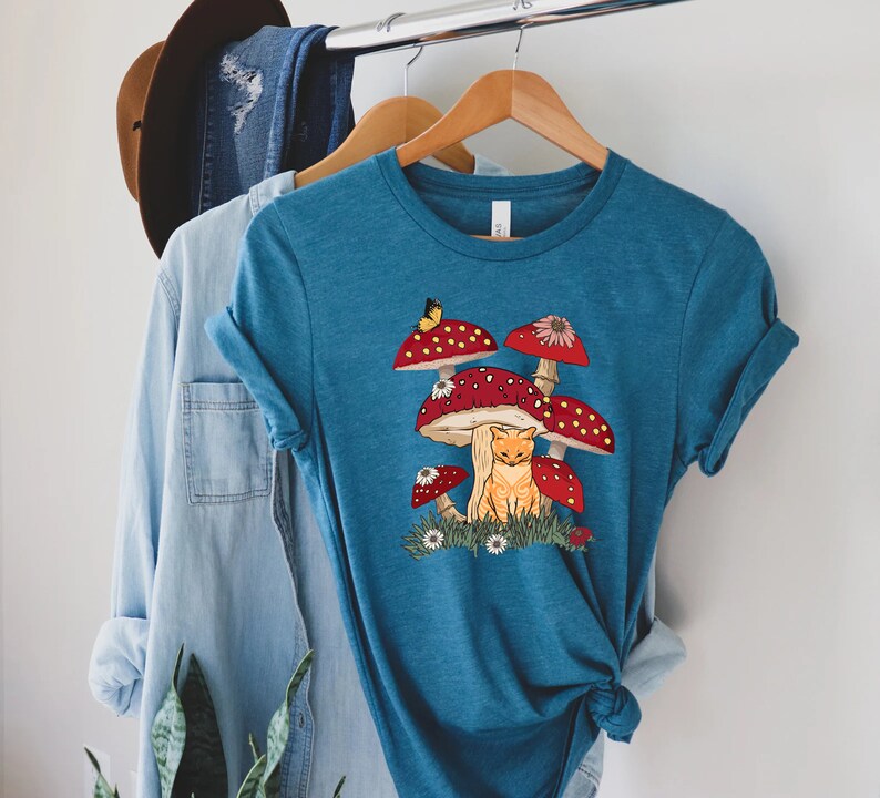 May include: A teal blue t-shirt with a graphic of a cat sitting under a cluster of red and white mushrooms. A yellow butterfly is flying above the mushrooms.