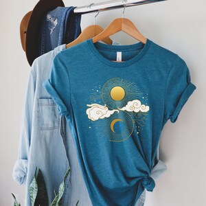 May include: A teal blue t-shirt with a gold sun and moon design. The design features a crescent moon, a sun with rays, and clouds.