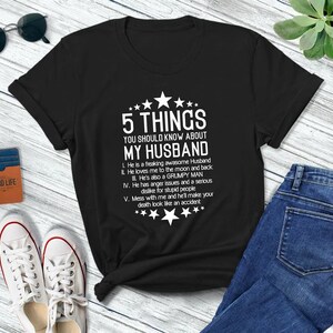 以下が含まれることがあります： 黒いTシャツに白い文字で「夫について知っておくべき5つのこと」と書かれています。テキストには、夫に関するユーモラスなポイントが5つリストされています。
