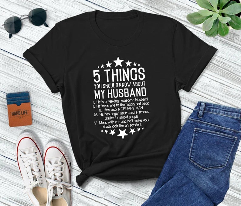 以下が含まれることがあります： 黒いTシャツに白い文字で「夫について知っておくべき5つのこと」と書かれています。テキストには、夫に関するユーモラスなポイントが5つリストされています。