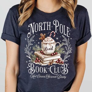 K&ouml;nnte beinhalten: Ein marineblaues T-Shirt mit einem wei&szlig;en Grafikdesign, das eine Tasse hei&szlig;er Schokolade mit einem Lebkuchenmann, einen Stapel B&uuml;cher und den Text "North Pole Book Club. Hot Cocoa Served Daily." zeigt.