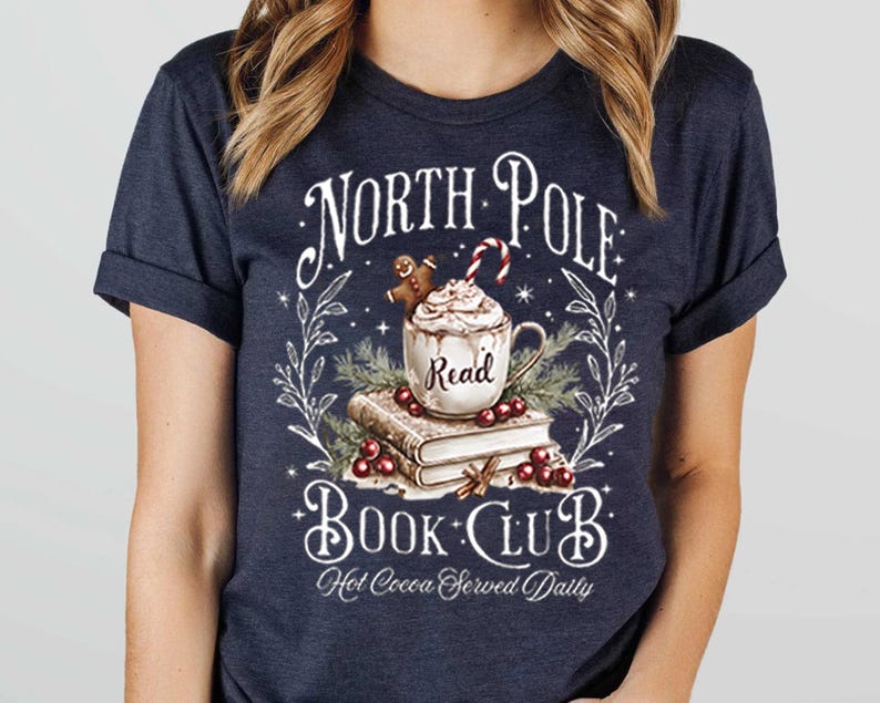 K&ouml;nnte beinhalten: Ein marineblaues T-Shirt mit einem wei&szlig;en Grafikdesign, das eine Tasse hei&szlig;er Schokolade mit einem Lebkuchenmann, einen Stapel B&uuml;cher und den Text "North Pole Book Club. Hot Cocoa Served Daily." zeigt.