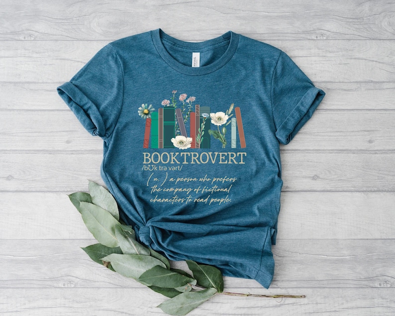 Puede incluir: Camiseta azul turquesa con un gr&aacute;fico de libros apilados con flores y el texto "BOOKTROVERT /b&uuml;k trə vərt/ (n.) a person who prefers the company of fictional characters to read people."