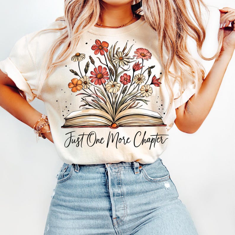以下が含まれることがあります： 花が咲いている開いた本が描かれたクリーム色のTシャツ。本の下に「Just One More Chapter」という文字が印刷されています。