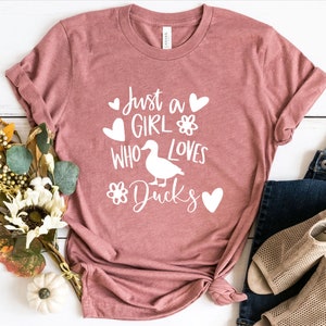 May include: A dusty rose t-shirt with the text "Just a Girl Who Loves Ducks" in white. The design includes a duck silhouette, hearts, and floral accents. The shirt is styled with blue jeans and beige sandals.