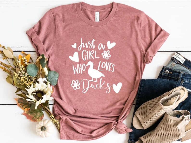 May include: A dusty rose t-shirt with the text "Just a Girl Who Loves Ducks" in white. The design includes a duck silhouette, hearts, and floral accents. The shirt is styled with blue jeans and beige sandals.