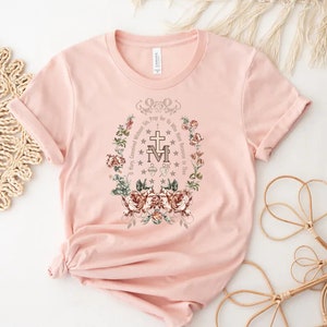 Op de afbeelding: Een roze T-shirt met een bloemenprint en de tekst "O Maria, ontvangen zonder zonde, bid voor ons die onze toevlucht tot u nemen".