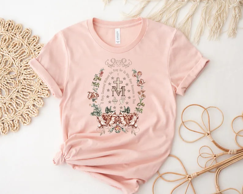Op de afbeelding: Een roze T-shirt met een bloemenprint en de tekst "O Maria, ontvangen zonder zonde, bid voor ons die onze toevlucht tot u nemen".