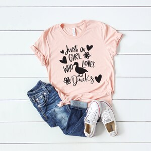 May include: A light pink t-shirt with the text "Just a GIRL who loves Ducks" in black, along with a duck graphic and heart and flower designs. The shirt is paired with blue jeans and white sneakers.